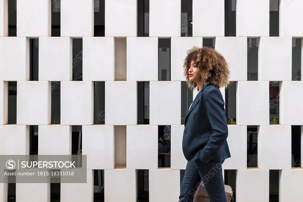 Afro female entrepreneur with hand in pocket walking by modern wall