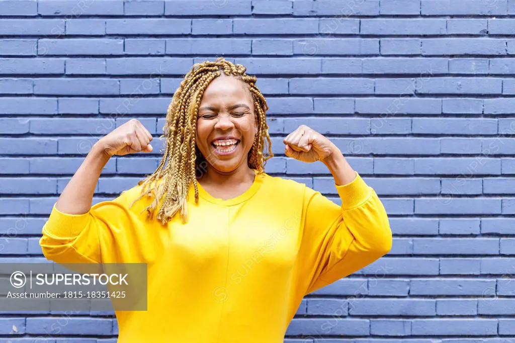 Cheerful plus size woman with eyes closed flexing muscles in front of brick wall