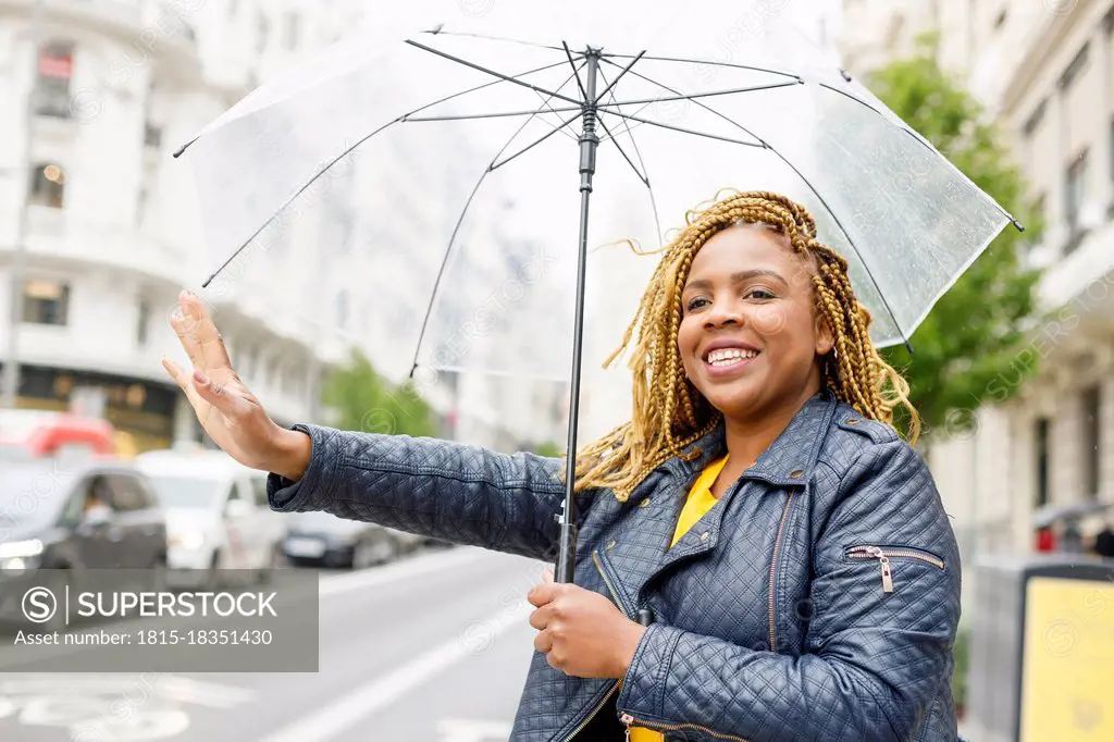 Happy woman with braids holding umbrella while hailing taxi in city