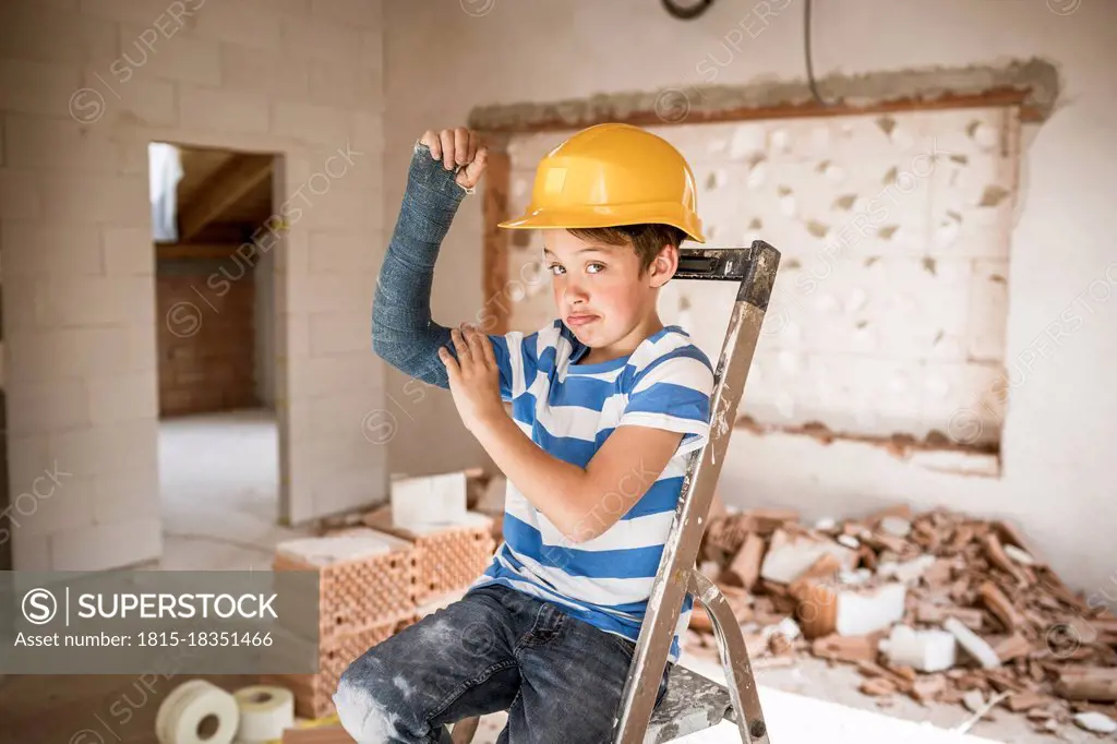 Boy flexing broken arm muscles while sitting on ladder during house renovation