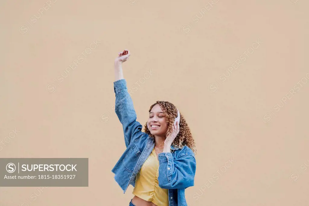 Woman with hand raised listening music on headphones by beige wall