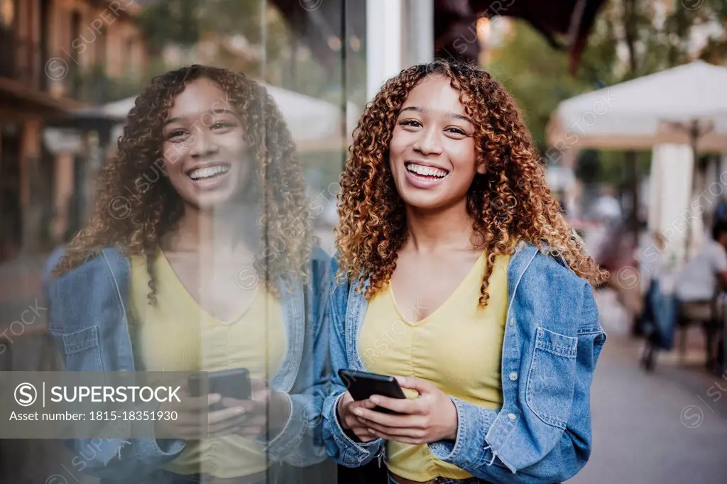 Smiling woman with smart phone leaning on glass wall