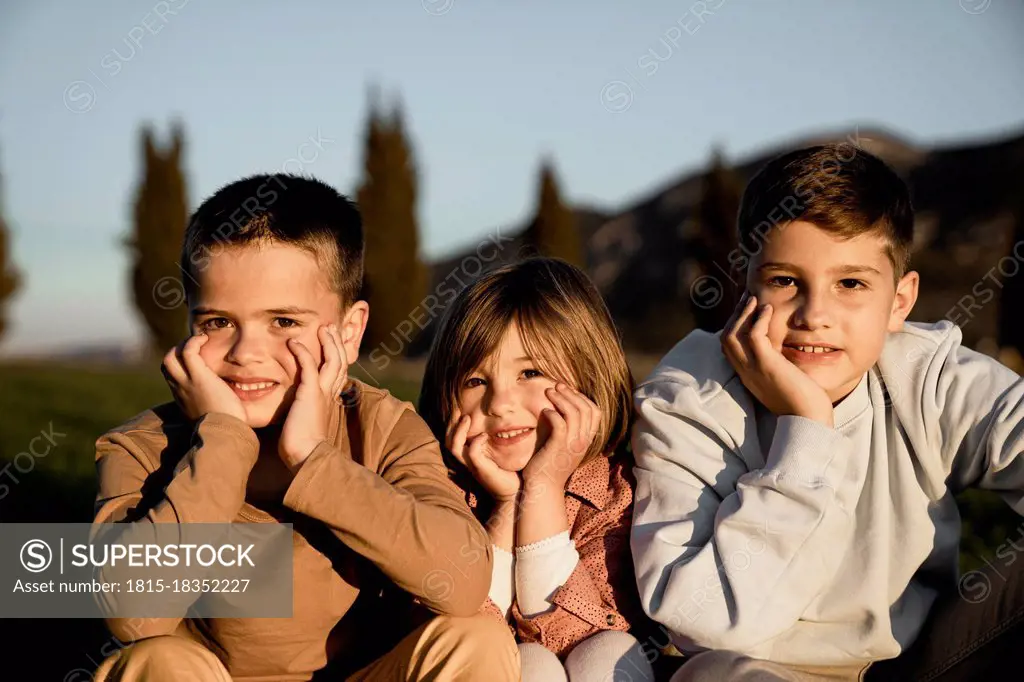 Smiling brothers and sister sitting with hand on chin during sunny day