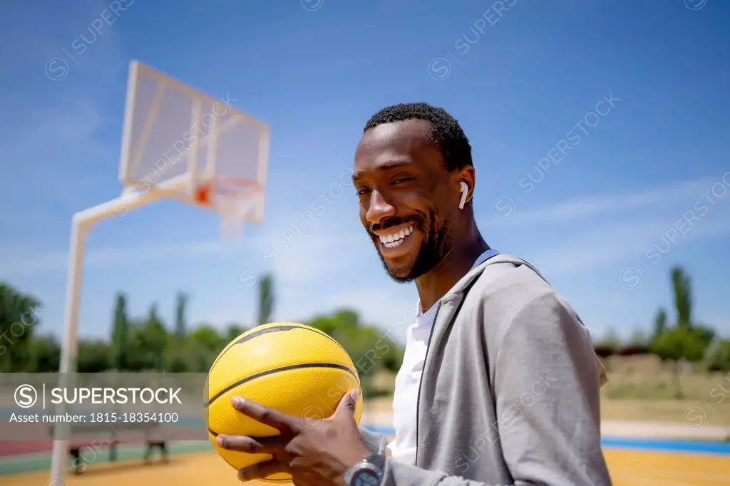 Smiling young man with wireless in-ear headphones holding basketball on sunny day