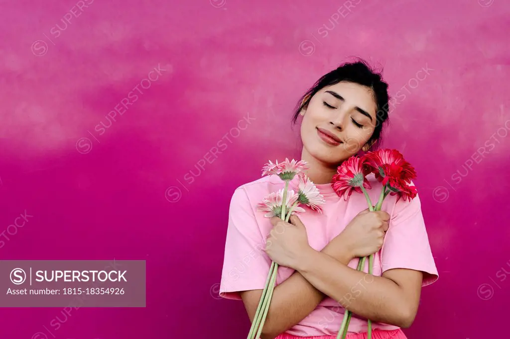 Young woman with arms crossed holding bunch of Gerbera daisies in front of pink wall