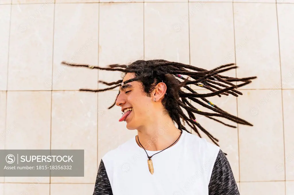 Man with dreadlocks shaking head while sticking out tongue in front of wall