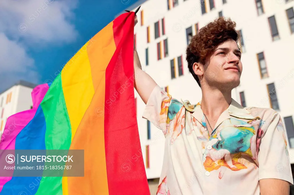 Young man looking away while blowing rainbow flag