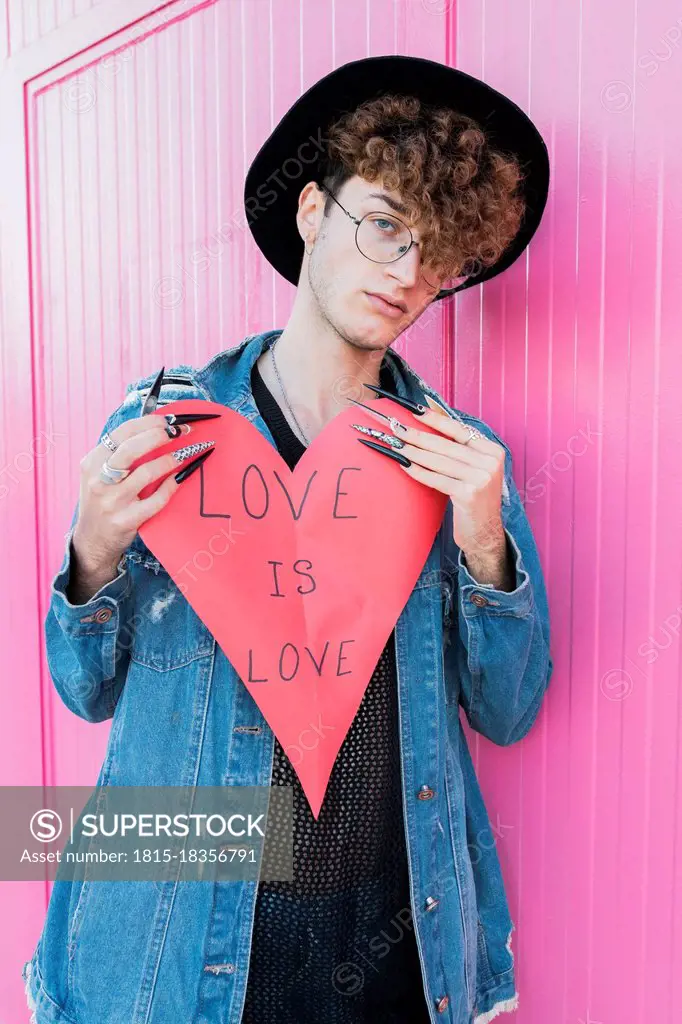 Young gay man with heart shape leaning on pink wall