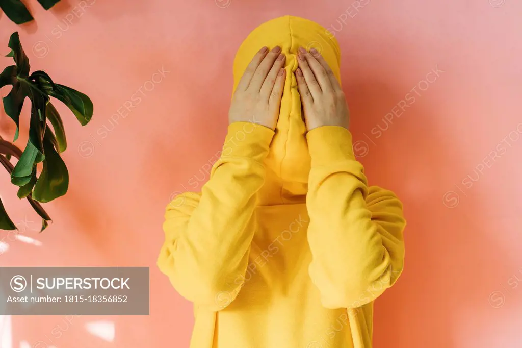 Young woman covering face with yellow hoodie in front of peach wall