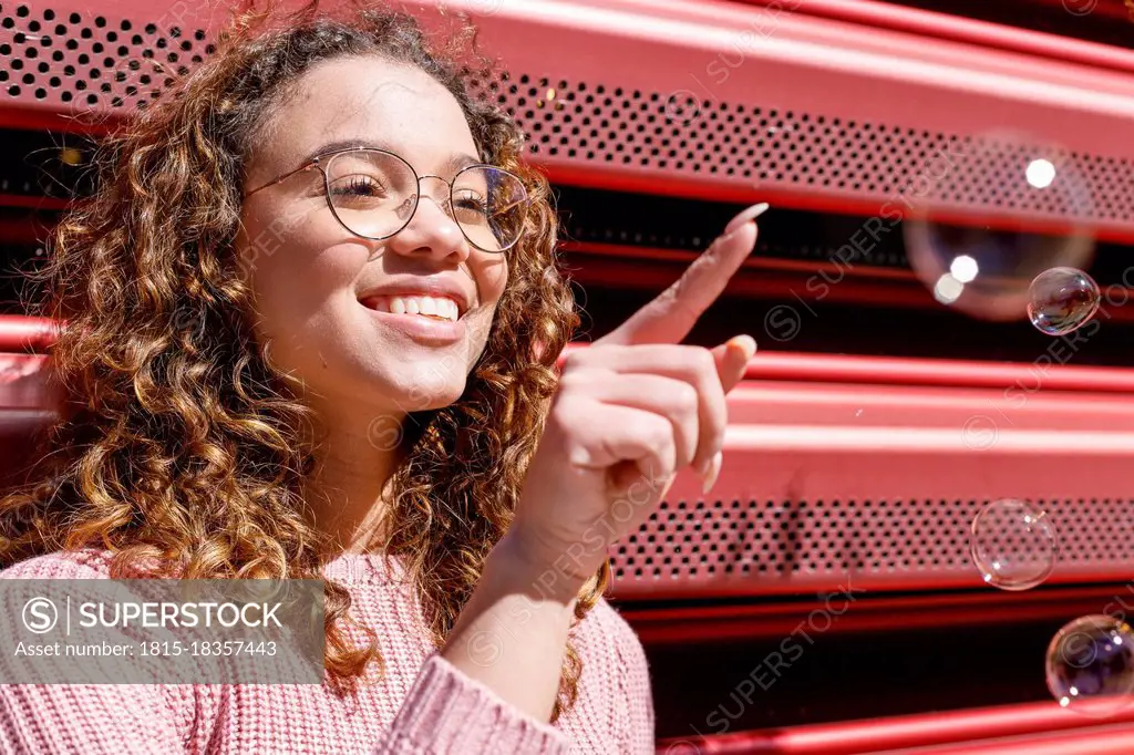 Happy young woman pointing at bubbles on sunny day