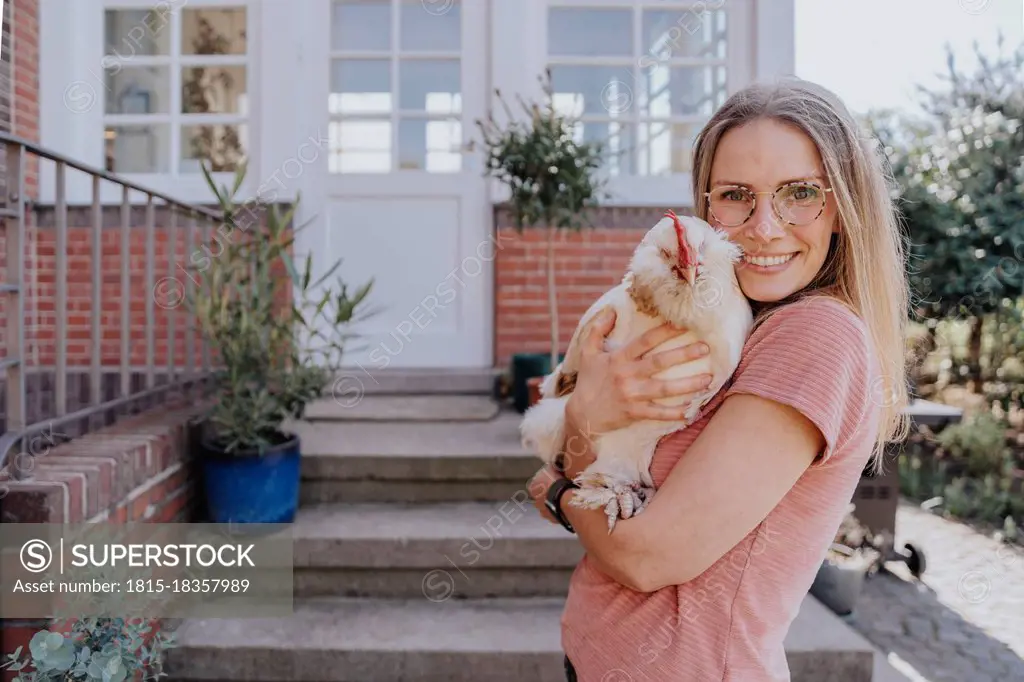 Smiling woman embracing chicken while standing in backyard