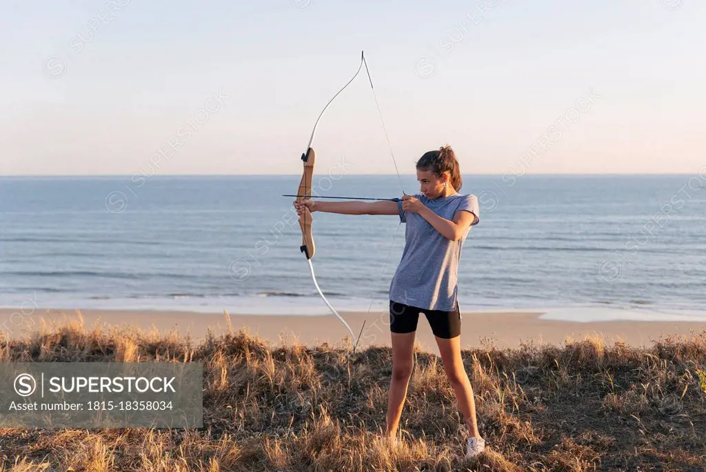 Archeress aiming with bow and arrow while standing on grass at beach