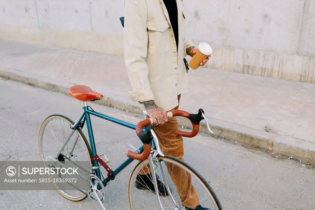 Man holding reusable coffee cup while wheeling bicycle on road