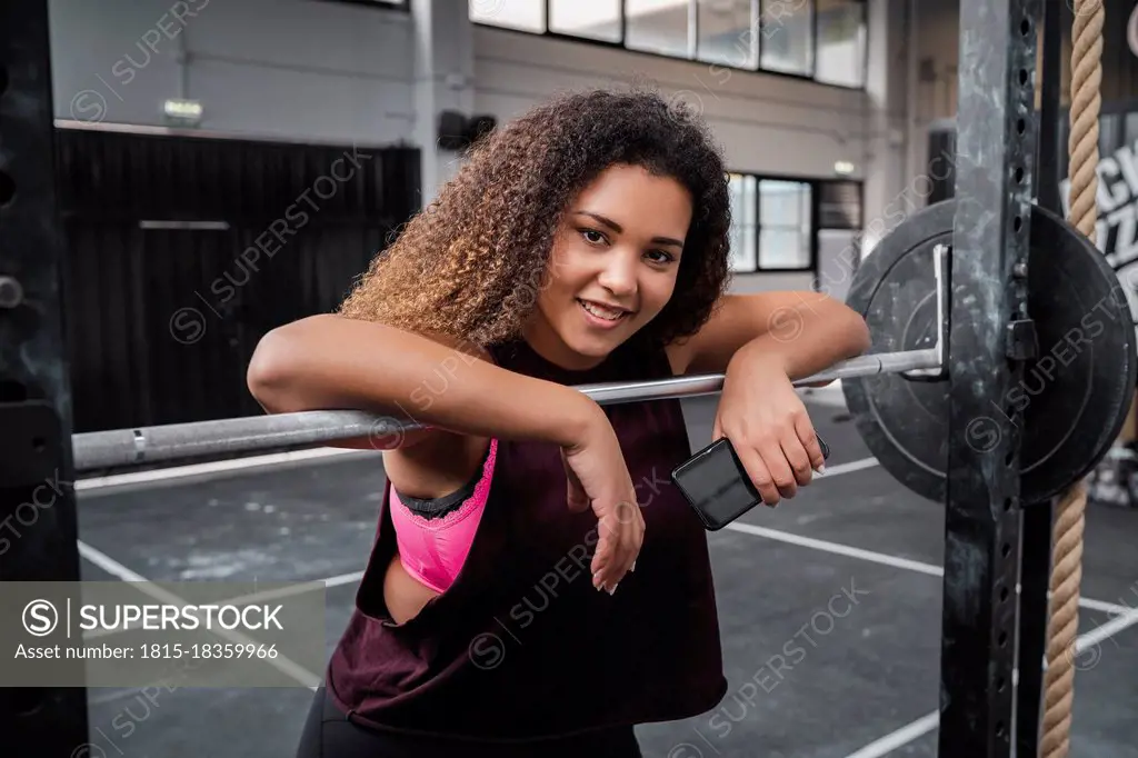 Smiling female athlete with mobile phone leaning on barbell at gym