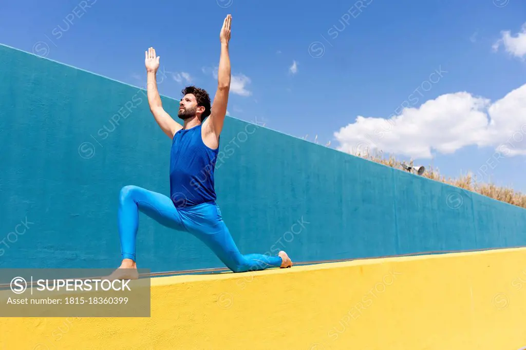 Man with arms raised practicing yoga on retaining wall during sunny day