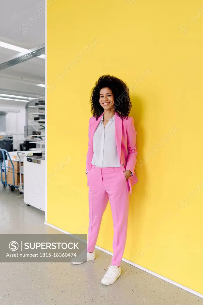 Smiling female entrepreneur with hands in pockets standing in front of yellow wall