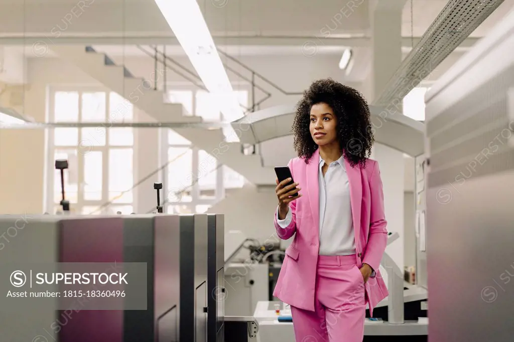 Female professional with mobile phone looking away while standing amidst machines in industry