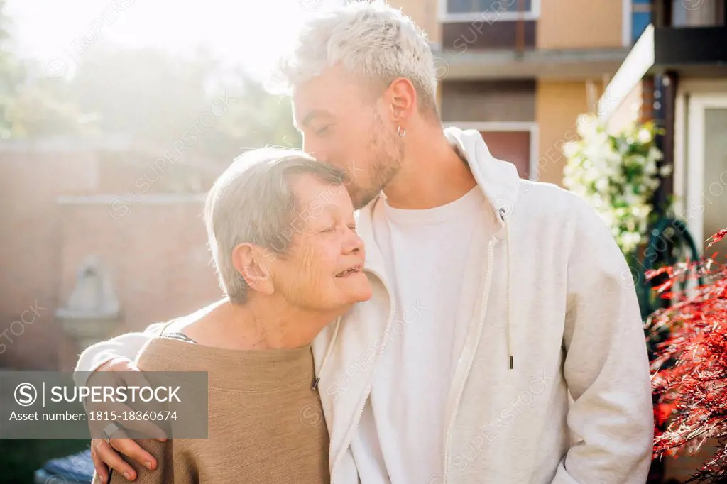 Young man kissing grandmother forehead while standing with arm around at backyard