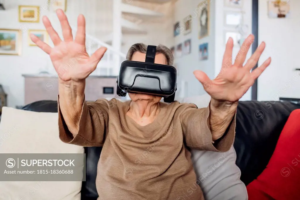 Senior woman wearing virtual reality simulator gesturing while siting on sofa in living room at home