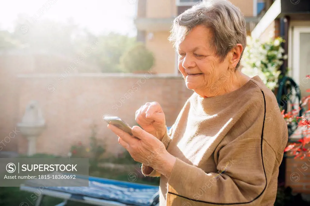 Smiling senior woman using smart phone while standing at backyard during sunny day