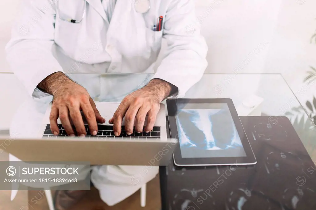 Senior male doctor working on laptop at desk in clinic