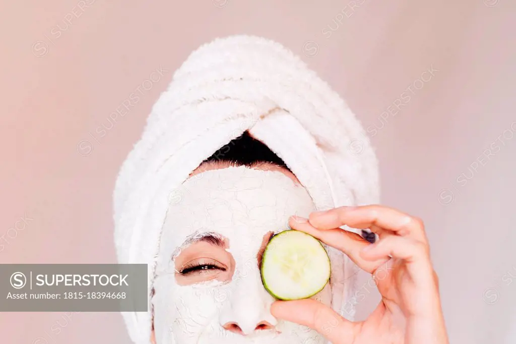 Young woman with beauty face mask covering eye with cucumber slice