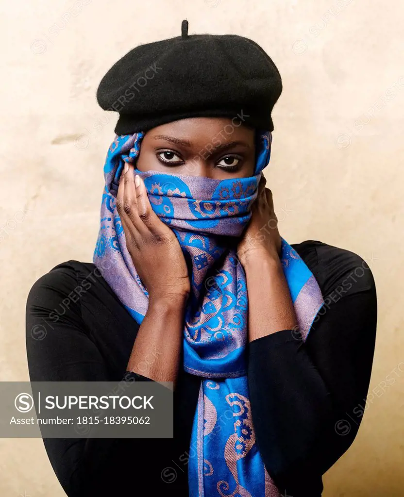 Young woman covering face with headscarf in front of beige wall