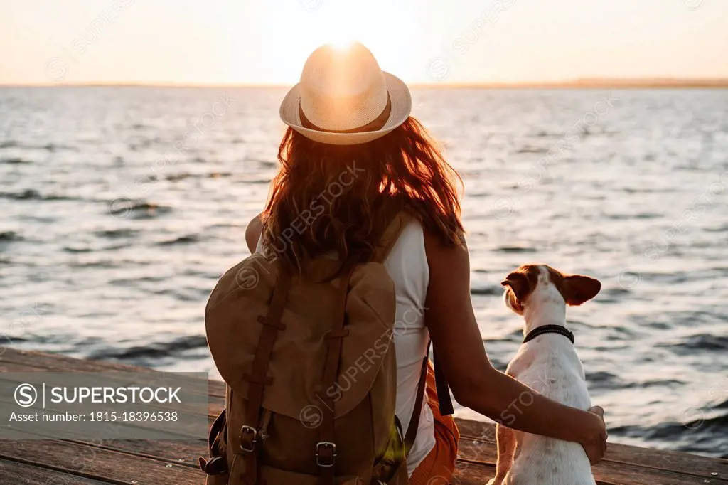 Woman with backpack and hat sitting by dog on pier