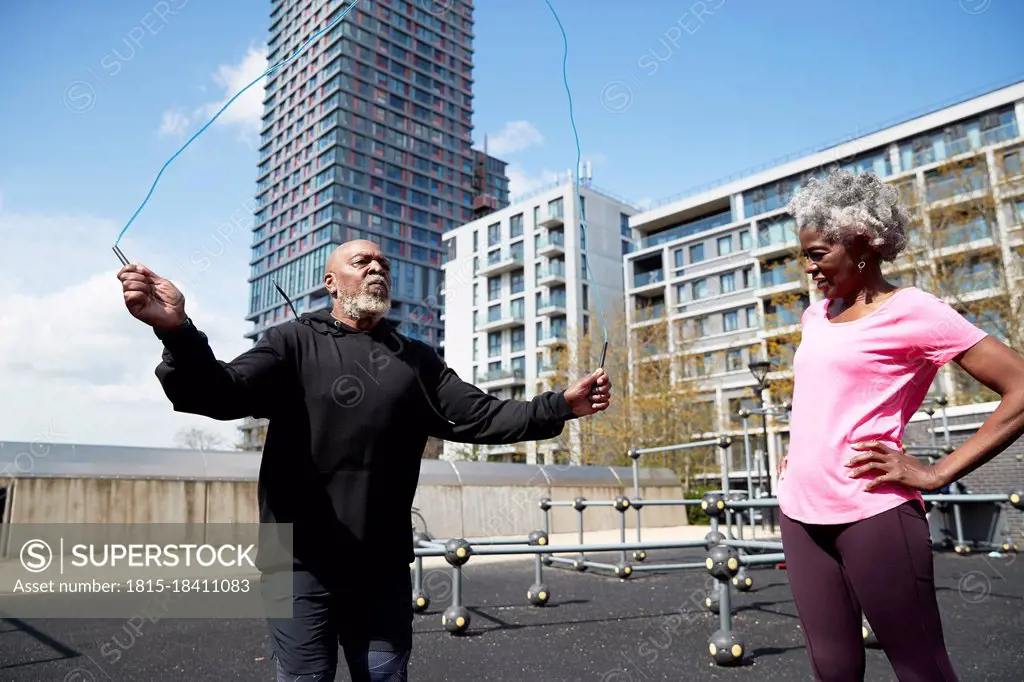 Woman with hands on hip standing by man skipping rope in park