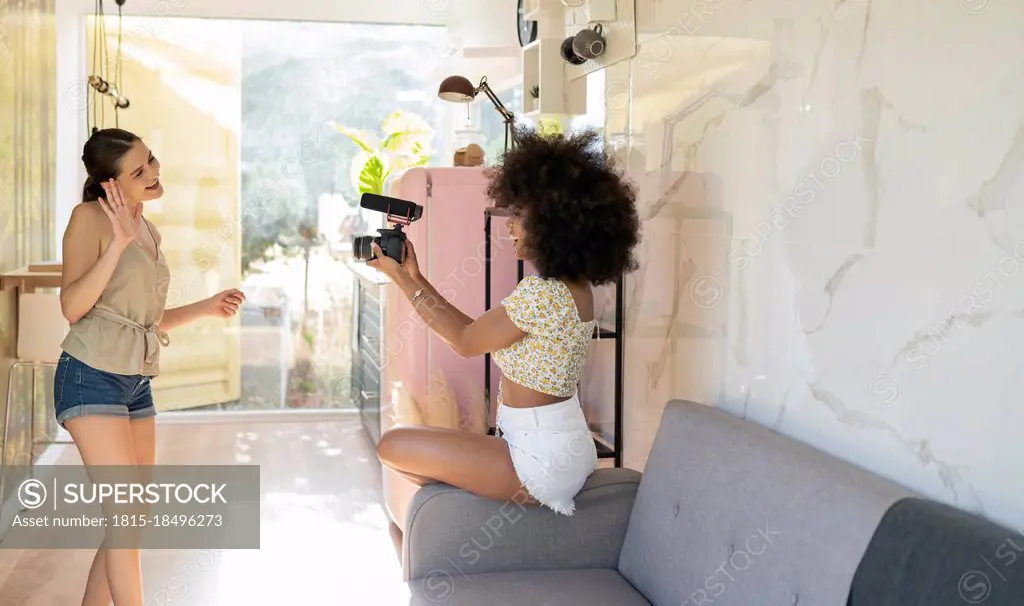 Young woman waving in camera held by afro female friend while filming at home