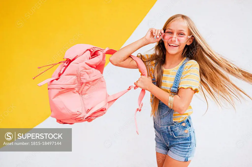 Happy girl with sunglasses and backpack spinning in front of wall