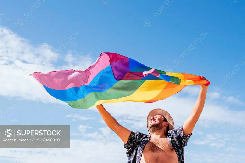 Cheerful gay man waving rainbow flag under sky on sunny day