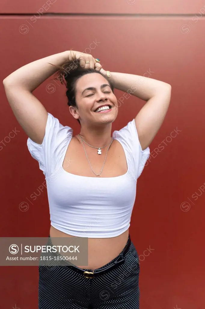 Cheerful young woman tying hair in front of brown wall