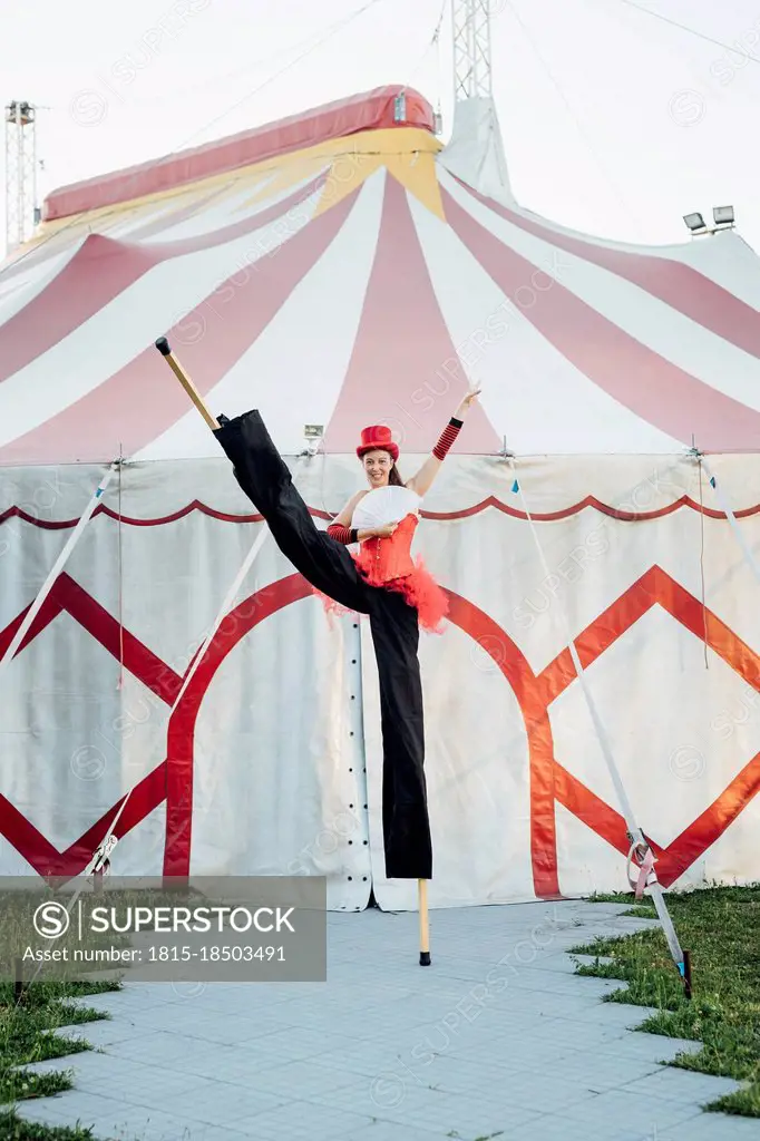 Female artist holding hand fan while standing with stilts in front of tent