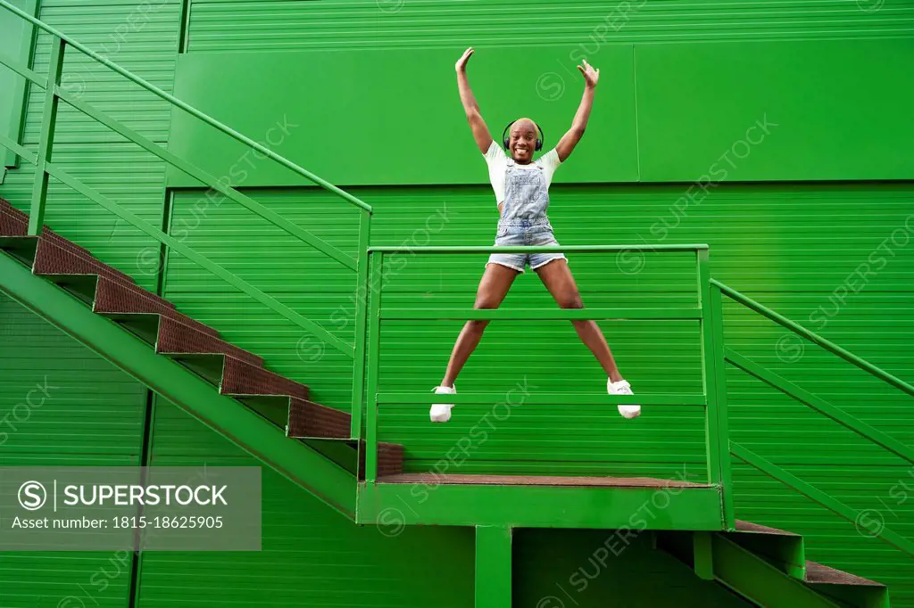 Happy woman with hands raised jumping while listening music on staircase in front of green wall