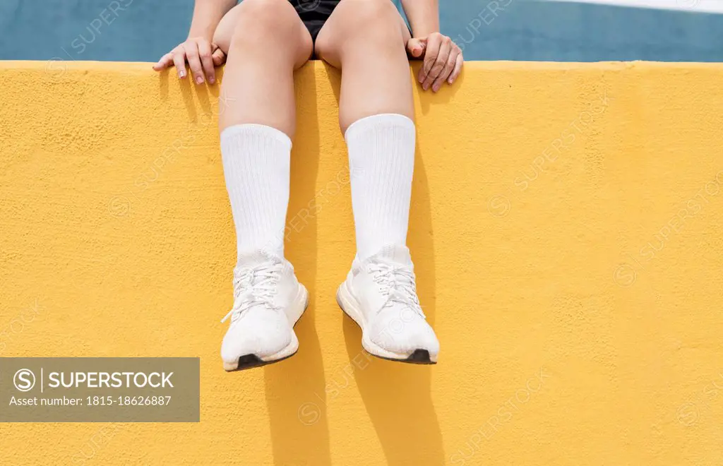 Woman wearing white sports shoes sitting on yellow retaining wall