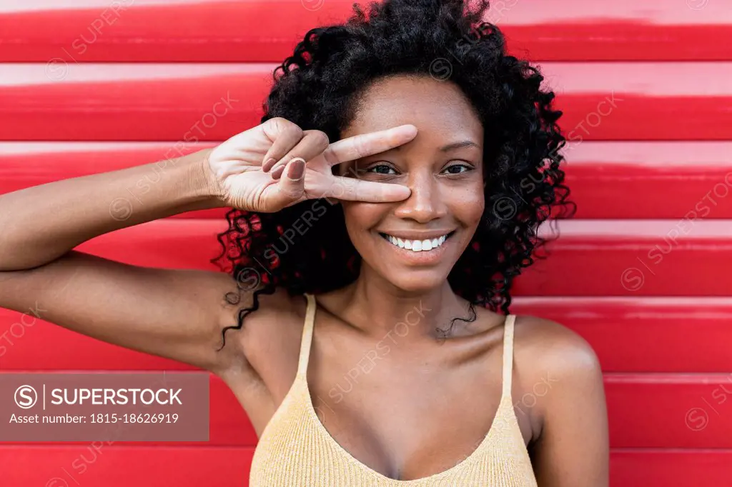 Smiling woman with curly hair gesturing peace sign in front of red shutter