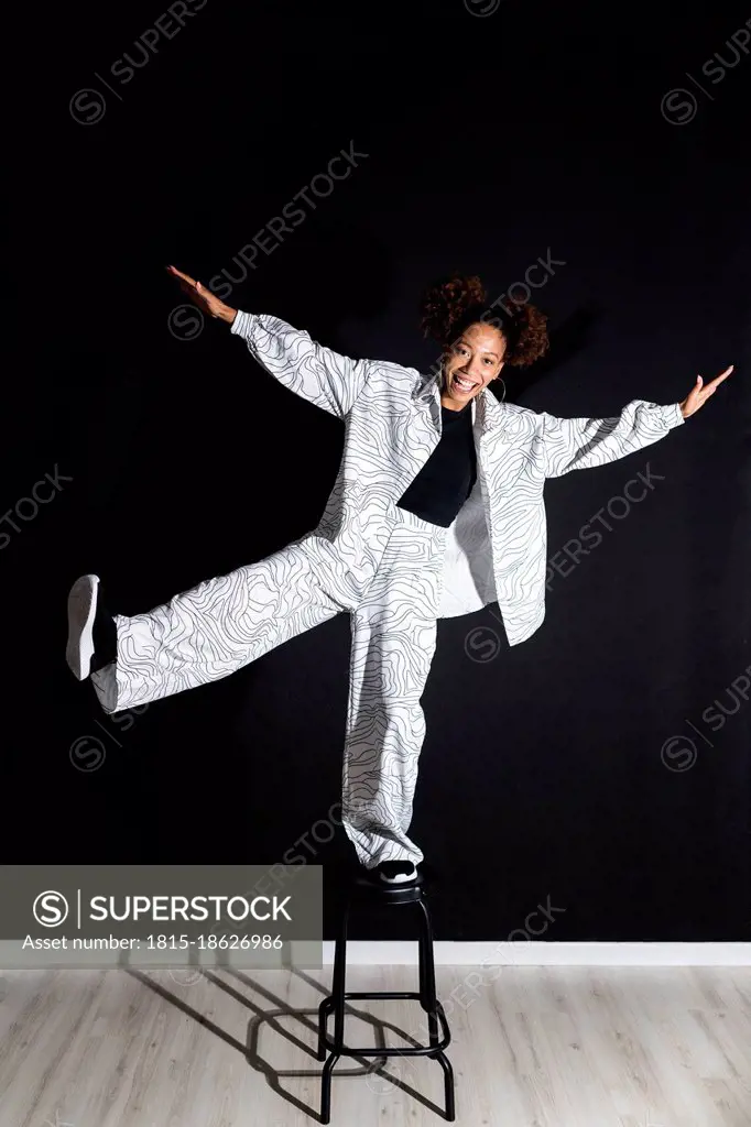 Happy fashionable woman balancing on stool in front of black wall