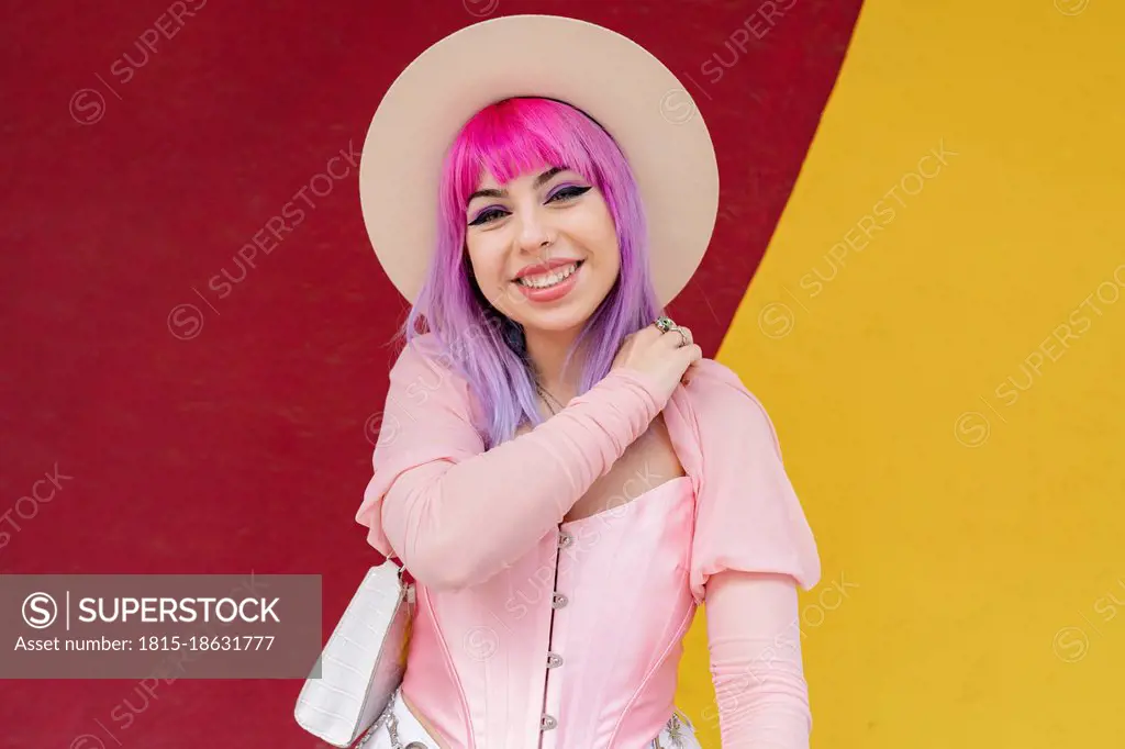 Fashionable young woman with dyed hair smiling in front of wall