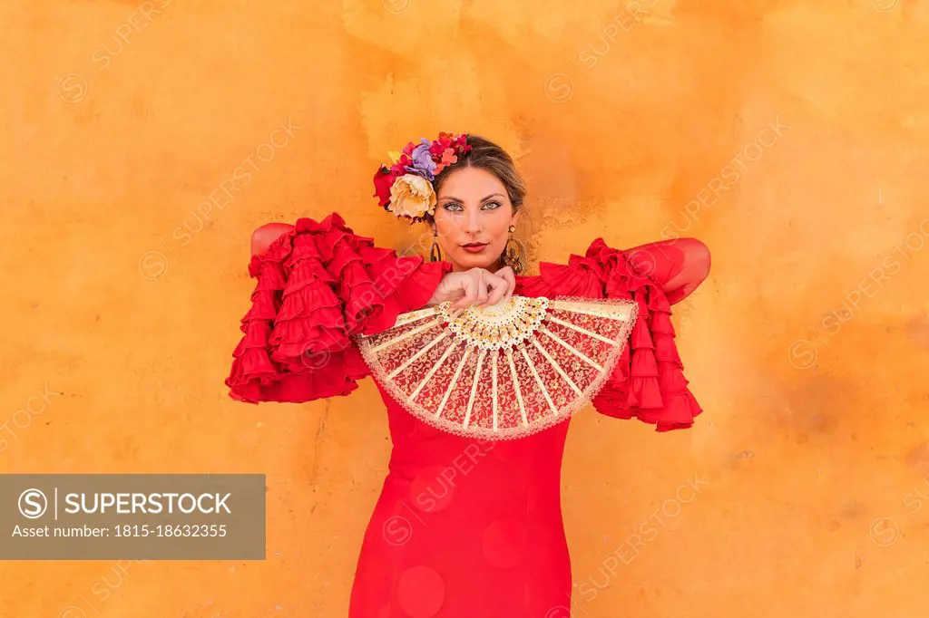 Female flamenco artist holding hand fan while standing in front of orange wall