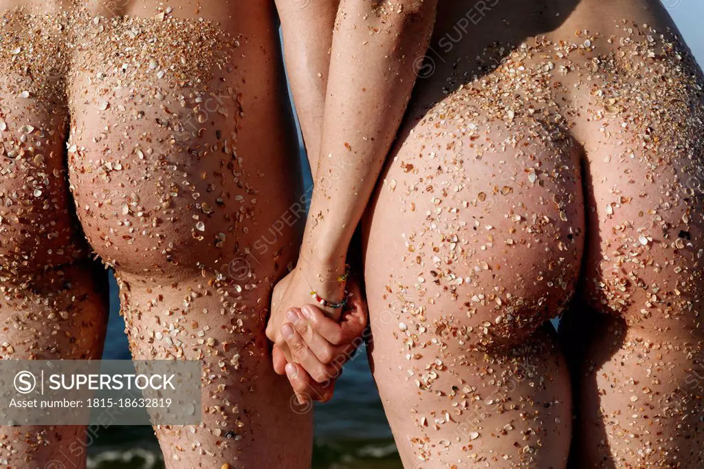 Naked couple with sand on buttocks holding hands while standing at beach