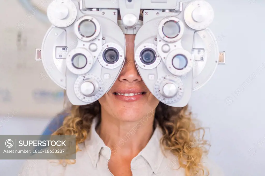 Smiling female patient examining eyesight through phoropter in medical clinic