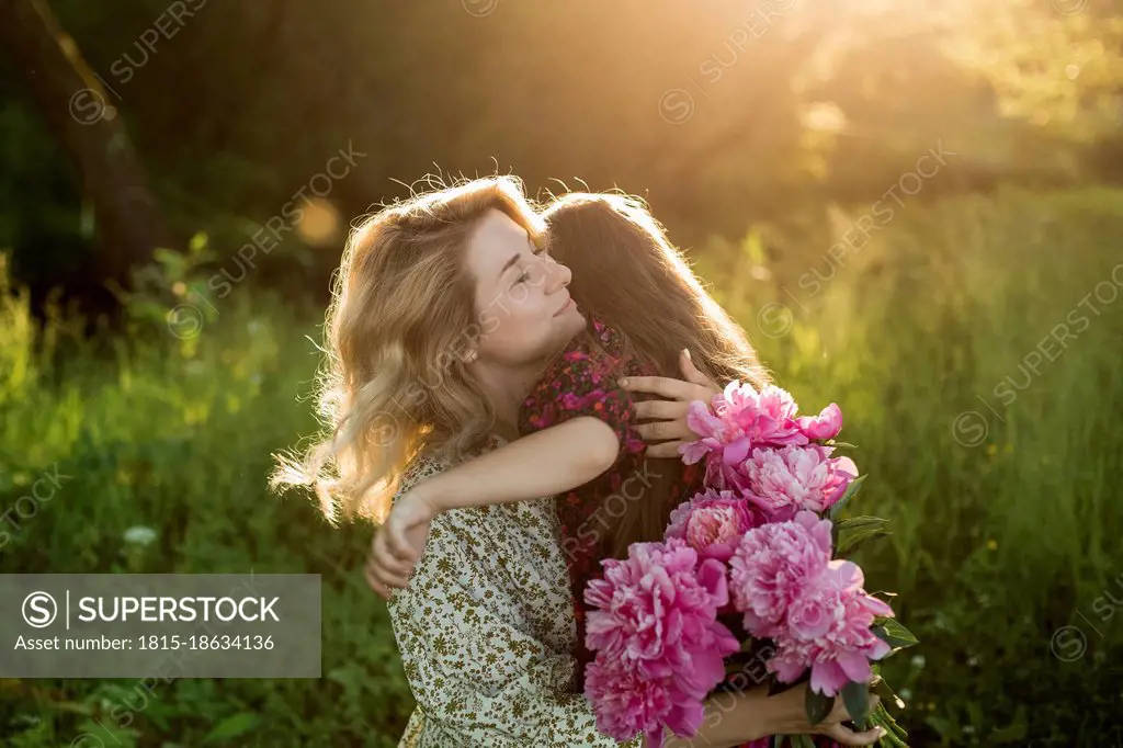 Woman with bunch of peonies embracing girl at meadow