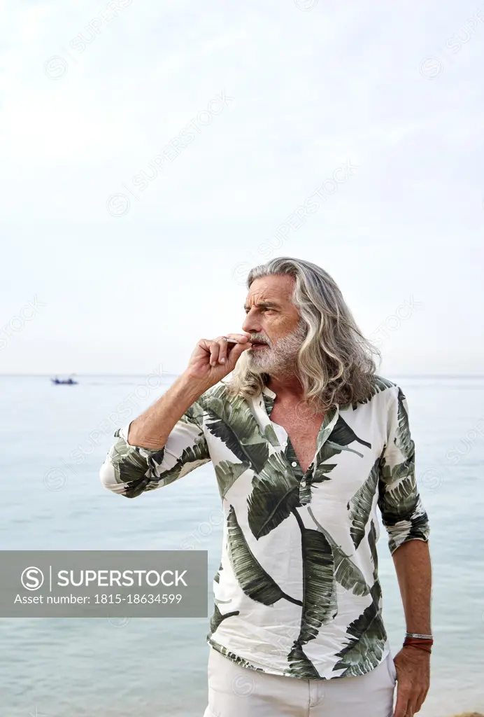 Mature man with gray hair smoking near sea at beach