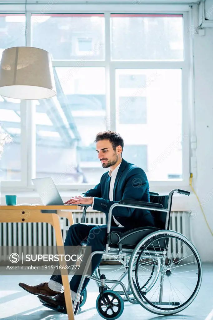 Young disabled male professional using laptop while sitting in wheelchair in office