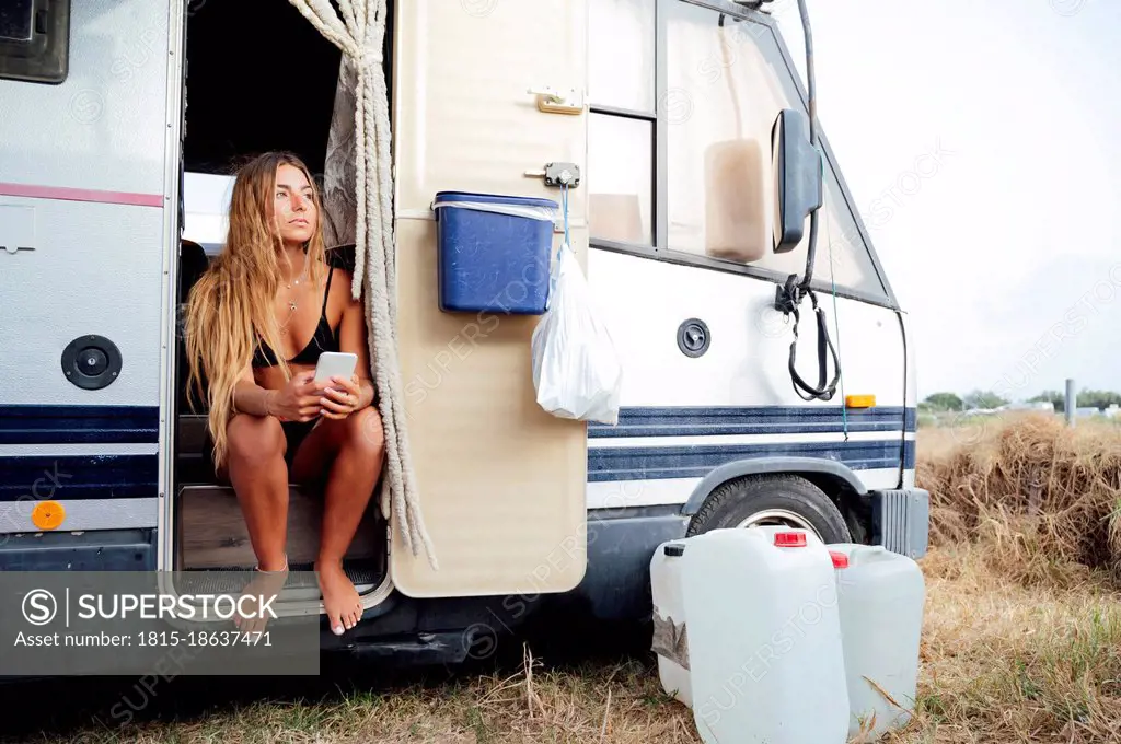 Young woman holding smart phone while sitting at doorway of camping van