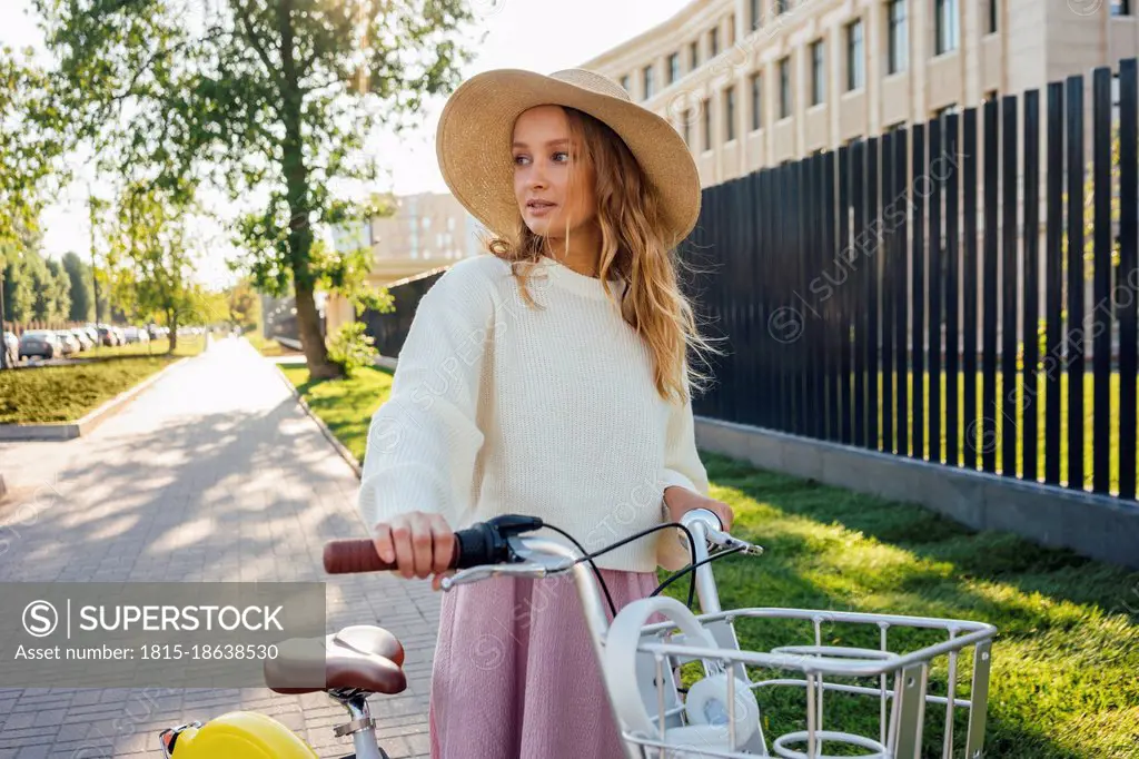 Young blond woman wearing hat standing with bicycle at park