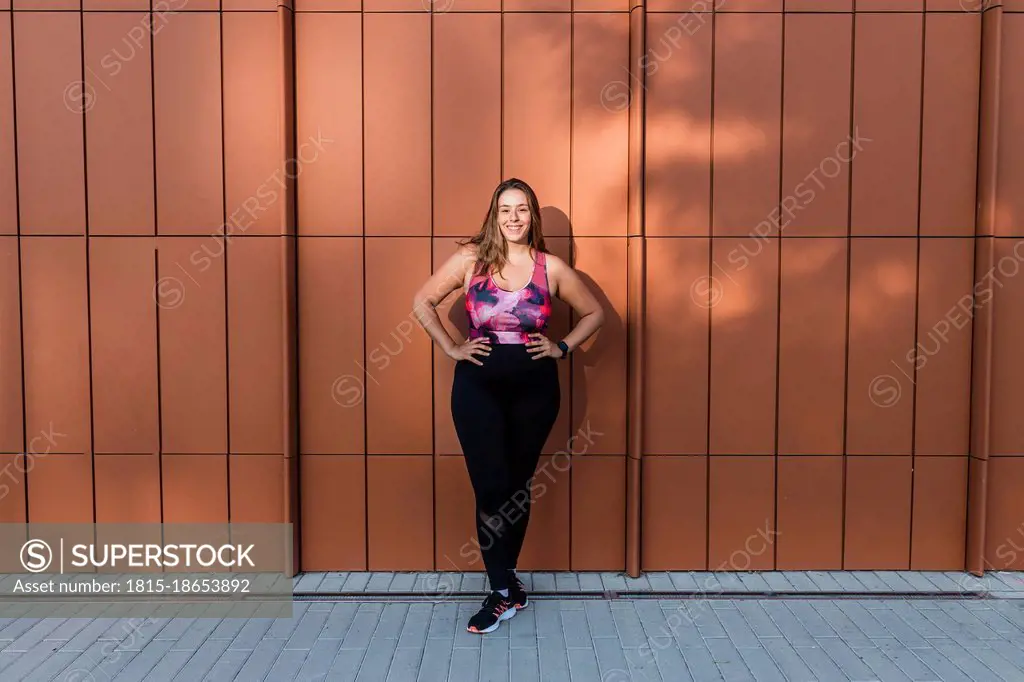 Smiling sportswoman with hand on hip standing in front of wall