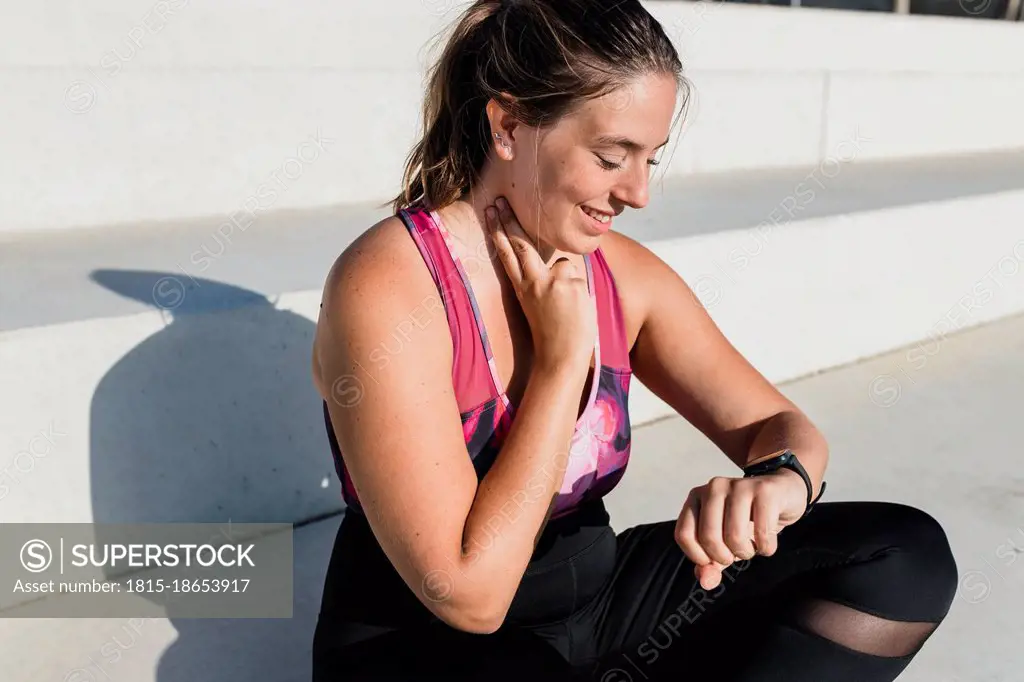 Smiling young woman looking at smart watch while checking pulse on neck during sunny day