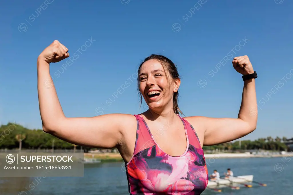 Sportswoman laughing while flexing muscles at riverbank